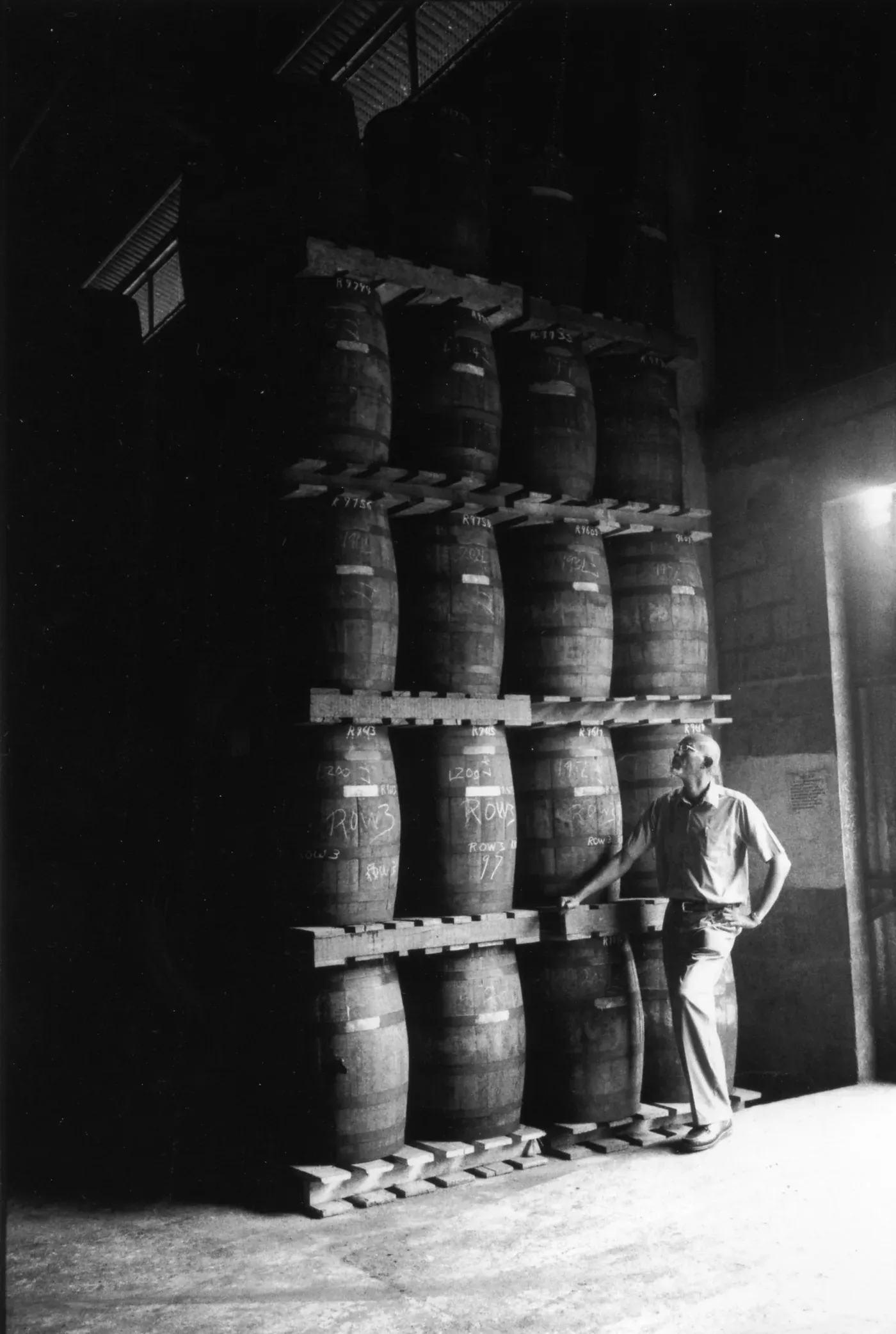 A man at the foot of a tall tower of stacked Caroni casks in the dark warehouse